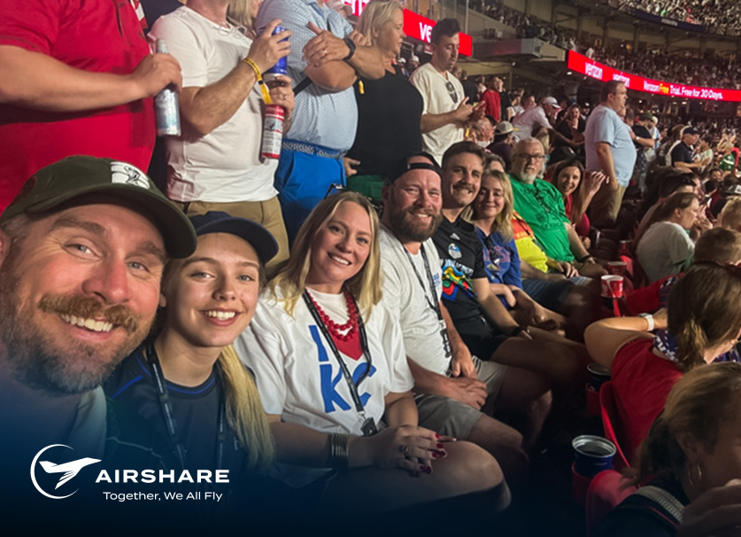 Group of Airshare employees seated together at a packed soccer stadium during a match, with the Airshare logo and tagline 'Together, We All Fly' displayed in the lower-left corner.